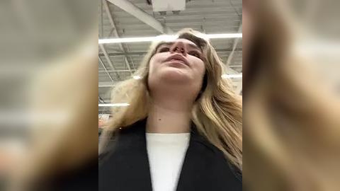 Media: Video of a Caucasian woman with long blonde hair, wearing a black jacket over a white shirt, tilted head back, smiling broadly, in an industrial setting with exposed pipes and ceiling.