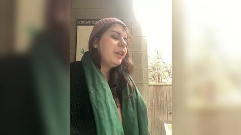 Media: Video of a young woman with light skin and long dark hair, wearing a red knit beanie, green scarf, and black sweater, standing outdoors near a wooden fence.