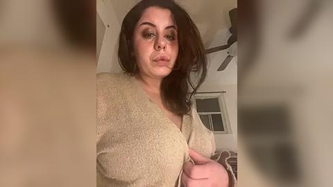 Media: Video of a curvy woman with medium skin tone, wearing a beige cardigan, standing indoors. Her dark hair is tousled, and she has a neutral facial expression.