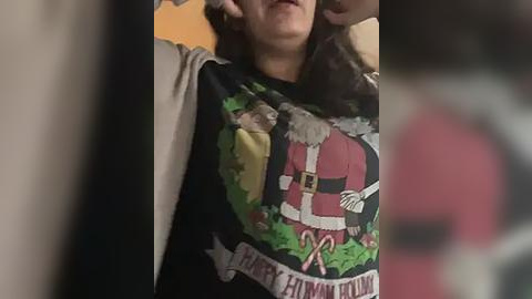 Media: Video of a woman wearing a black \"Santa Claus\" T-shirt with a cartoon Santa, candy cane, and greenery; blurred background, dim lighting.