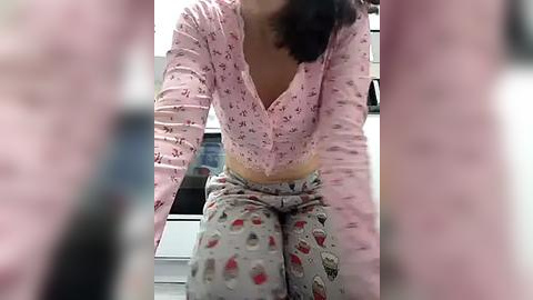 Media: Video of a woman with long dark hair, wearing a pink floral top and patterned pajama pants, seated indoors.