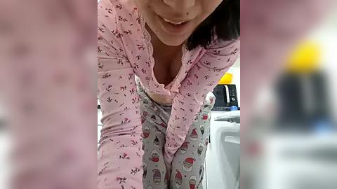 Media: A video of a woman with fair skin and shoulder-length dark hair, wearing a pink floral nightgown and pajama pants, leaning over a white sink, smiling. Background shows a blurred kitchen scene.