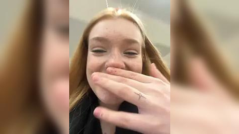 Media: A video of a young woman with long red hair, fair skin, and a freckled face, covering her mouth with her hands, smiling. The background is blurred, suggesting an indoor setting.