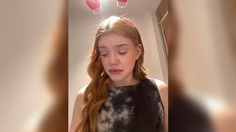 Media: Video of a young Caucasian girl with long, wavy, auburn hair wearing a black tie-dye shirt, bunny ears, and closed eyes in a bathroom with beige walls and a framed picture in the background.