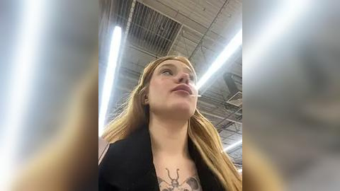 Media: Video of a young woman with long, straight red hair, wearing a black coat and a gray shirt, with a detailed tattoo on her chest, standing in a dimly lit, industrial warehouse with high ceilings and exposed pipes.