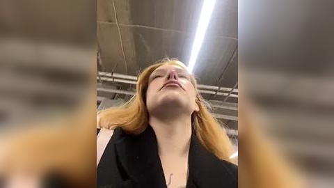 Media: Video of a smiling woman with long, straight, light-brown hair, wearing a black jacket, standing in an industrial room with a high ceiling, fluorescent lights, and visible pipes.