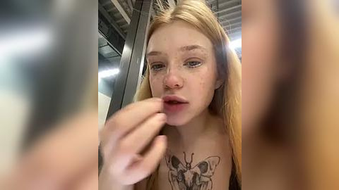 Media: Video of a young, fair-skinned woman with long blonde hair, wearing a black top, applying lipstick. She has a detailed butterfly tattoo on her chest. Background shows industrial ceiling and blurred faces.