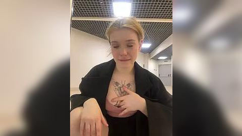 Media: Video of a young woman with blonde hair, wearing a black jacket, touching her exposed, tattooed breasts in a dimly lit hallway with a grid ceiling and fluorescent lights.