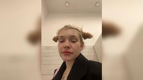 Media: Video of a young woman with blonde hair in twin buns, wearing a black blazer, standing in a white-tiled bathroom with recessed ceiling lights. Her expression is neutral.