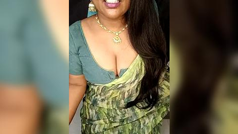 Media: Video of a South Asian woman with medium brown skin, wearing a teal top with a plunging neckline, green and gold sari, and gold necklace, smiling, blurred background.