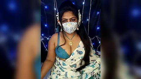 Media: Video of a woman with dark skin, wearing a teal bra, white sari with blue floral print, white face mask, and ornate headpiece, sitting in a dark room with blue fairy lights in the background.