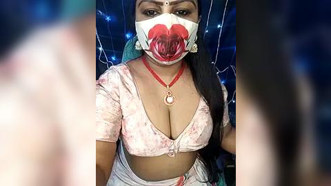 Media: Video of a South Asian woman with medium skin tone, wearing a floral-patterned crop top revealing ample cleavage, red mask, and red necklace, in a dimly lit room with blue fairy lights.