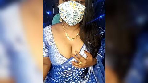 Media: Video of a South Asian woman with dark skin, long dark hair, wearing a blue floral-patterned dress, large gold necklace, and a white floral-patterned face mask, covering her mouth and nose.