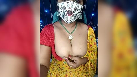 Media: Video of a woman with medium brown skin, wearing a yellow saree with floral pattern, exposing cleavage, and a green mask with intricate designs.