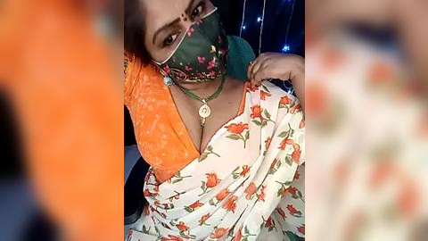 Media: Video of a woman with medium brown skin, wearing a floral saree, orange blouse, green mask, and a necklace, taken indoors.