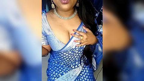 Media: Video of a South Asian woman with medium brown skin, wearing a blue sari with a deep V-neck, revealing ample cleavage. She has long, straight black hair, adorned with large gold earrings and a matching necklace. Her manicured nails are painted red.