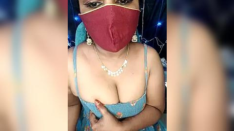 Media: Video of a South Asian woman with medium skin tone, wearing a turquoise saree with gold floral embroidery, red face mask, and gold earrings. She has medium-sized breasts, visible cleavage. Background is blurred, featuring blue fairy lights.