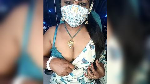 Media: Video of a dark-skinned woman with medium breasts, wearing a blue and white floral saree, gold jewelry, and a white mask, against a blurred background.