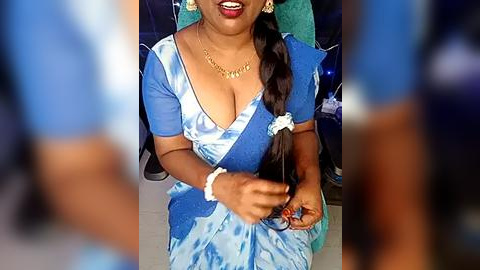 Media: Video of a South Asian woman with medium brown skin, smiling, wearing a blue sari with a plunging neckline, gold jewelry, and holding a bottle.