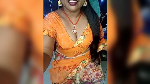 Media: Video of a South Asian woman with medium-dark skin, wearing an orange saree with floral patterns and a deep V-neck, large gold earrings, and a red necklace, smiling.