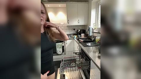 Media: Video of a woman in a black tank top and shorts, standing in a modern kitchen with stainless steel appliances and white cabinets, looking into the dishwasher.