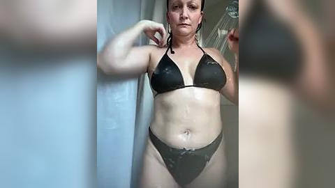 Media: Video of a muscular, fair-skinned woman with a wet, black bikini, posing confidently in front of a mirror. She has a toned physique with visible abs and medium-sized breasts. Background includes a blurred bathroom setting.