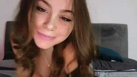 Media: Video of a young woman with long, wavy brown hair, light skin, and subtle makeup, smiling gently, leaning forward slightly. She wears a light-colored top and is indoors on a bed with gray and teal pillows.