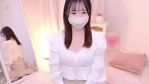 Media: Video of an Asian woman with light skin, long brown hair, wearing a white blouse, face mask, and kneeling in a soft-lit, pastel-toned room with a bed and mirror.