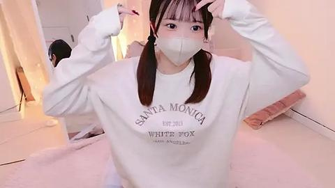 Media: Video of an East Asian woman in a white Santa Monica sweatshirt, wearing a face mask, adjusting her hair in a cozy, softly lit room with beige walls.