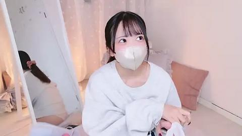 Media: Video of a young Asian woman with a white face mask, wearing a gray sweater, sitting on a bed in a softly lit, peach-toned room with a mirror reflecting her profile.