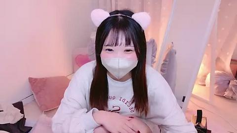 Media: Video of an East Asian woman with long black hair, wearing a white long-sleeve shirt, a white face mask, and cute white bear ears. She sits in a cozy bedroom with soft lighting, plush pillows, and a mirror.