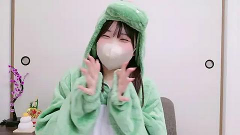 Media: Video of an Asian woman with straight black hair, wearing a green plush frog onesie, white mask, and oversized frog hands, indoors with white walls and a purple orchid plant in the background.