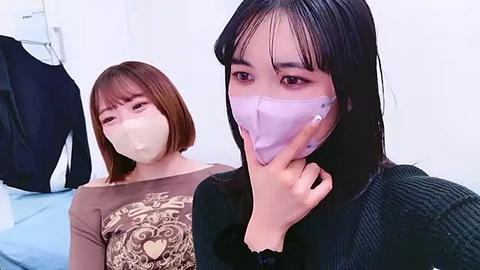 Media: Video of two East Asian women wearing surgical masks indoors. One with shoulder-length brown hair, another with straight black hair. Background shows white walls and a black jacket.