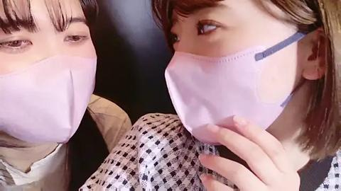 Media: Video of two Asian women with light skin, wearing pastel pink surgical masks, gazing closely at each other. One has straight brown hair, the other has bangs. They wear patterned blouses. Background is dark.