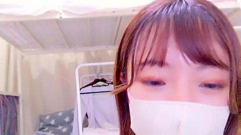 Media: Video of a young woman with straight brown hair, wearing a white surgical mask, standing in a hospital room with beige walls, a bed, and a chair.