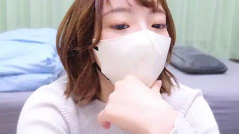 Media: Video of an Asian woman with shoulder-length brown hair, wearing a white face mask, white sweater, and light makeup, standing in a bedroom with light green curtains and a blue bedspread.