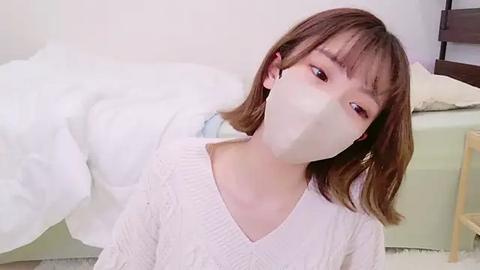 Media: Video of an East Asian woman with shoulder-length brown hair, wearing a white knit sweater and a face mask, lying on a white bed in a minimalist room.