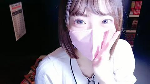 Media: Video of a young Asian woman with short brown hair, wearing a white mask, standing indoors with a dark background. She has a playful, curious expression, with one hand covering her mouth.