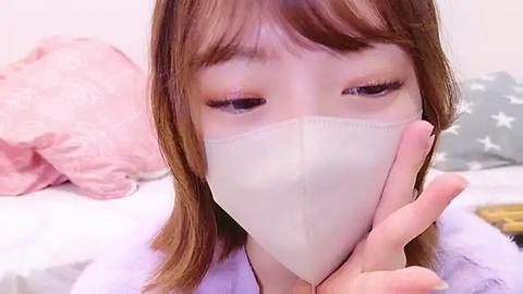 Media: Video of a young Asian woman with light skin and straight brown hair, wearing a white medical mask, making a peace sign with her right hand. Background shows a pink pillow and a star-patterned cushion.