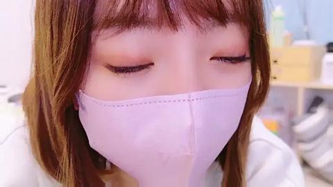 Media: A close-up video of a young Asian woman with straight, shoulder-length brown hair and bangs, wearing a white surgical mask, closed eyes, and a white jacket. The blurred background includes a wooden dresser with various objects.