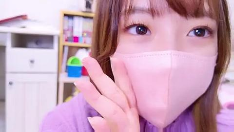 Media: Video of a young woman with light skin and brown hair, wearing a pink face mask, touching her nose, standing in a cluttered room with white furniture and colorful buckets.