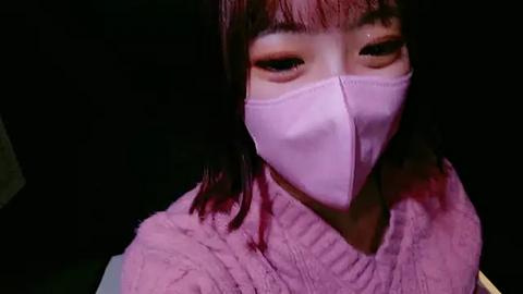 Media: Video of a young East Asian woman with straight, shoulder-length black hair, wearing a pink face mask and a light pink sweater, against a dark background.