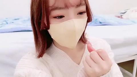 Media: Video of an East Asian woman with straight, shoulder-length red hair, wearing a beige face mask, a white knit sweater, and a white long-sleeve shirt. She sits on a white bed with blue sheets in a softly lit room.