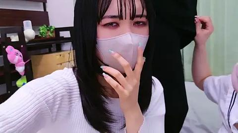 Media: Video of an Asian woman with long black hair, wearing a white sweater, white face mask, and light makeup, in a room with a wooden dresser, pink plush toy, and green curtains.