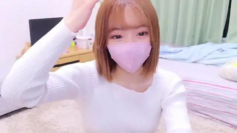 Media: Video of an Asian woman with straight, shoulder-length light brown hair, wearing a white ribbed sweater, pink face mask, and a light green curtain in the background.