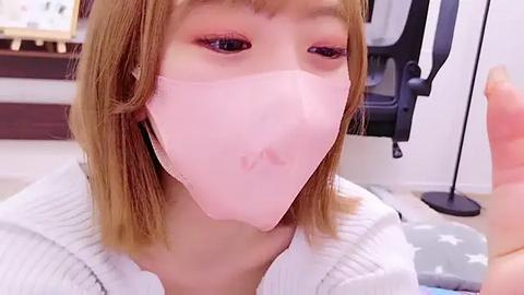 Media: Video of an East Asian woman with light skin, straight shoulder-length blonde hair, wearing a pink surgical mask and white ribbed sweater, sitting indoors with a modern room background.