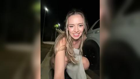 Media: A video of a topless blonde woman with wet hair, smiling, crouching by a parked car at night, wearing a white towel.