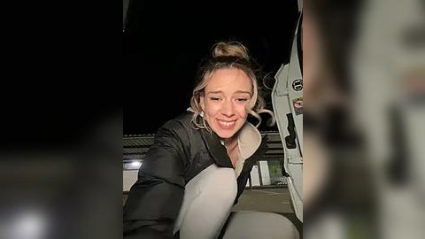 Media: Video of a young woman with blonde hair in a bun, wearing a black jacket, smiling and leaning on a white van at night.