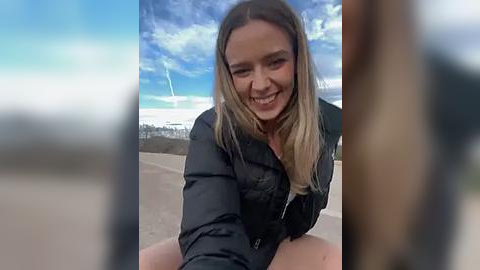 Media: Video of a smiling, light-skinned woman with long, straight blonde hair, wearing a black jacket, seated outdoors on a paved surface, with a clear blue sky and distant buildings visible in the background.