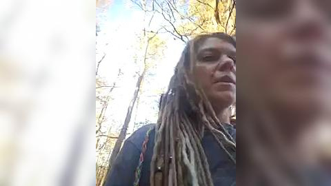 Media: Video of a middle-aged woman with long dreadlocks and a dark jacket standing in a forest. Sunlight filters through the tree branches, creating a warm, natural ambiance.